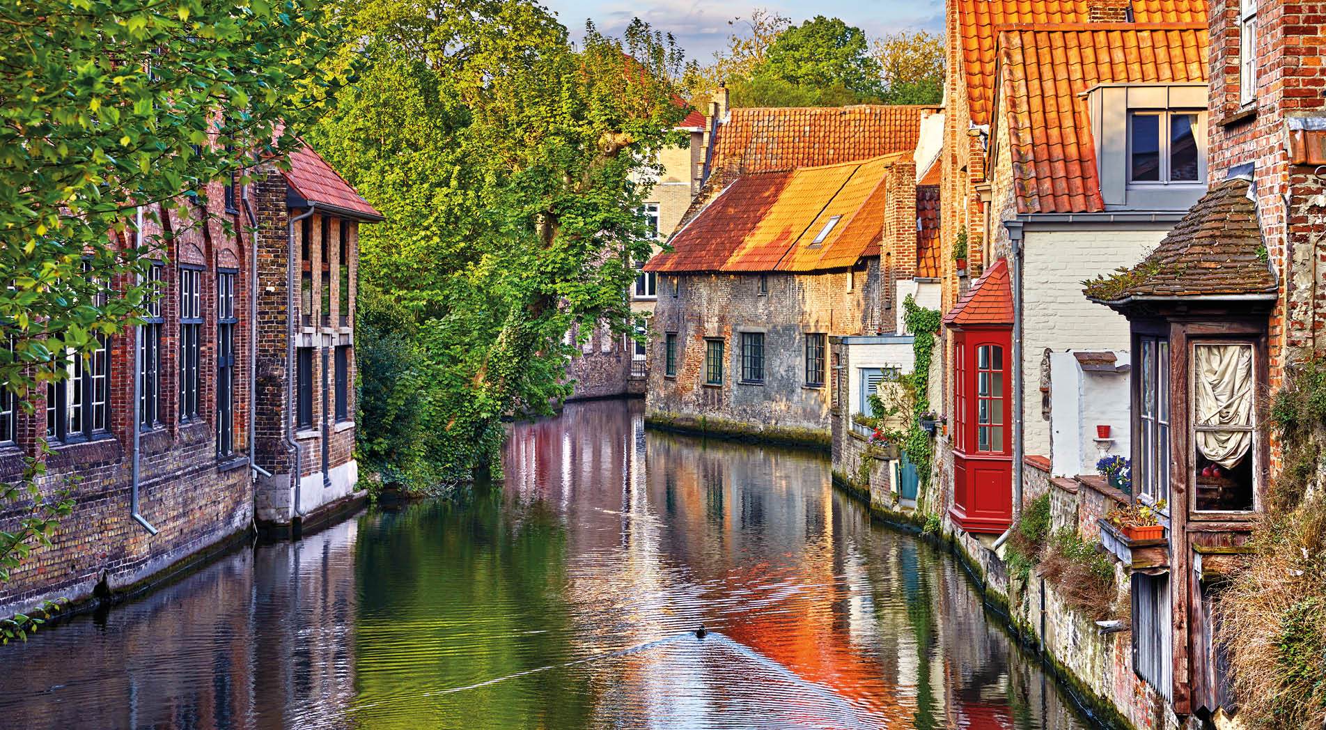 Bruges, Belgium. Medieval ancient houses made of old bricks at water channel with boats in old town. Summer sunset with sunshine and green trees. Picturesque landscape.
