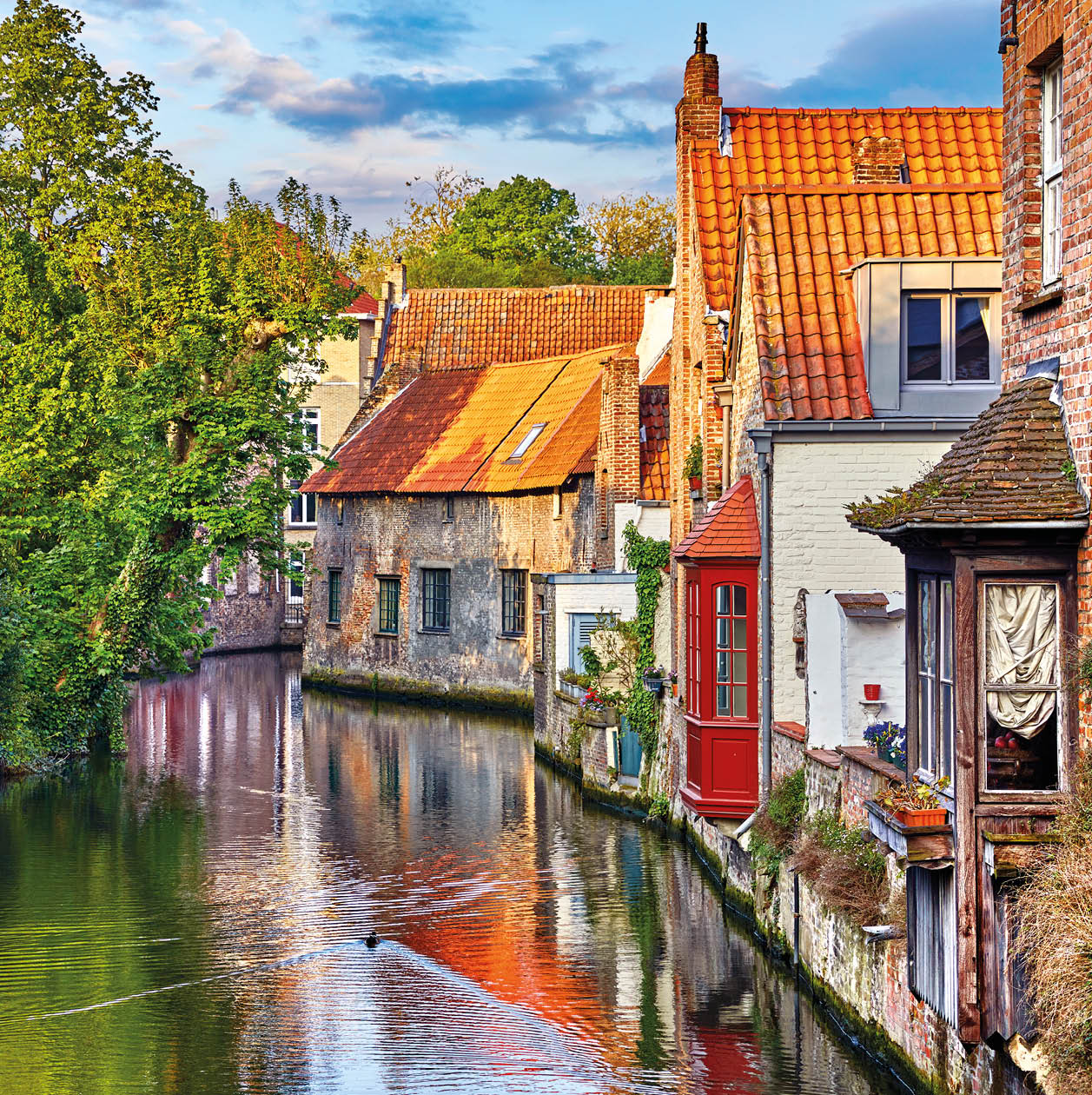 Bruges, Belgium. Medieval ancient houses made of old bricks at water channel with boats in old town. Summer sunset with sunshine and green trees. Picturesque landscape.