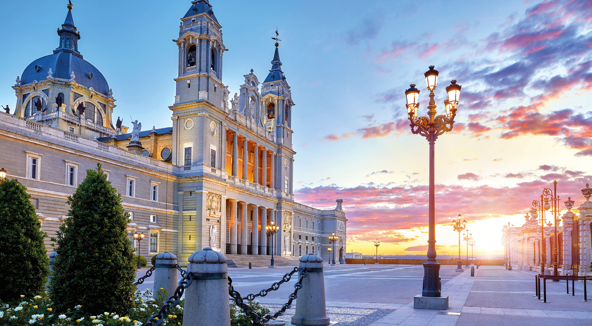 Madrid, Spain. Cathedral Santa Maria la Real de la Almudena at Plaza de la Armeria. Famous landmark with sunset sun, flowers and green bush. Street lamps with illumination and picturesque sky
