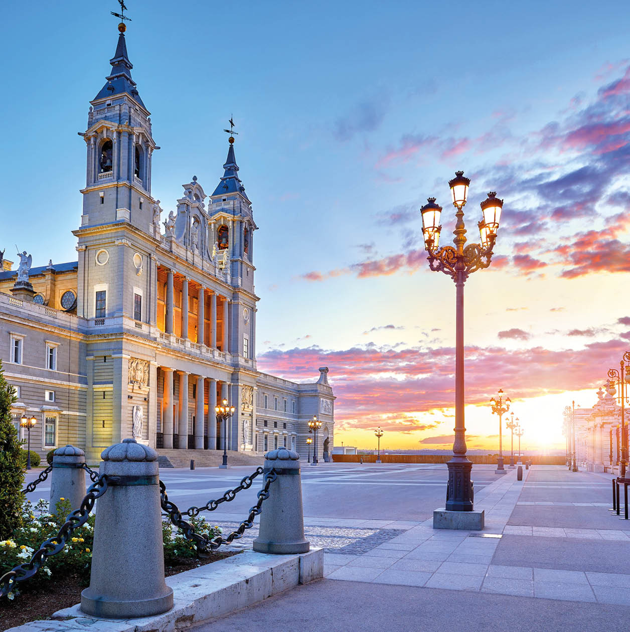 Madrid, Spain. Cathedral Santa Maria la Real de la Almudena at Plaza de la Armeria. Famous landmark with sunset sun, flowers and green bush. Street lamps with illumination and picturesque sky