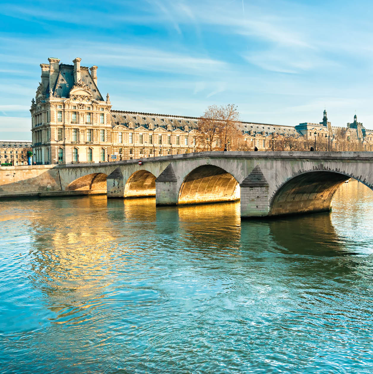 The Louvre art museum, seen from across the Seine River. The Pont Royal bridge crosses the Seine on the left and leads to the Louvre.  Both the bridge and the Louvre building are made of tan-colored stones.  The sky is blue and streaked with white wispy clouds.