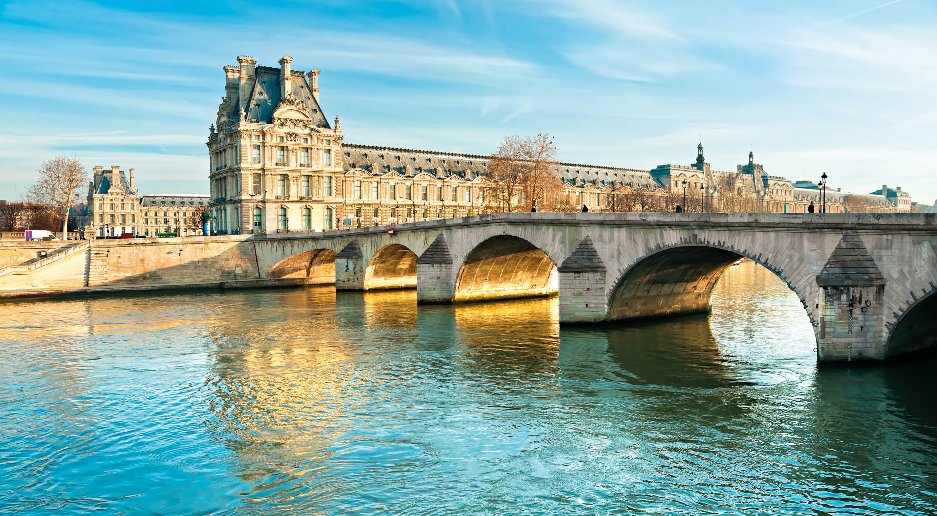 The Louvre art museum, seen from across the Seine River. The Pont Royal bridge crosses the Seine on the left and leads to the Louvre.  Both the bridge and the Louvre building are made of tan-colored stones.  The sky is blue and streaked with white wispy clouds.
