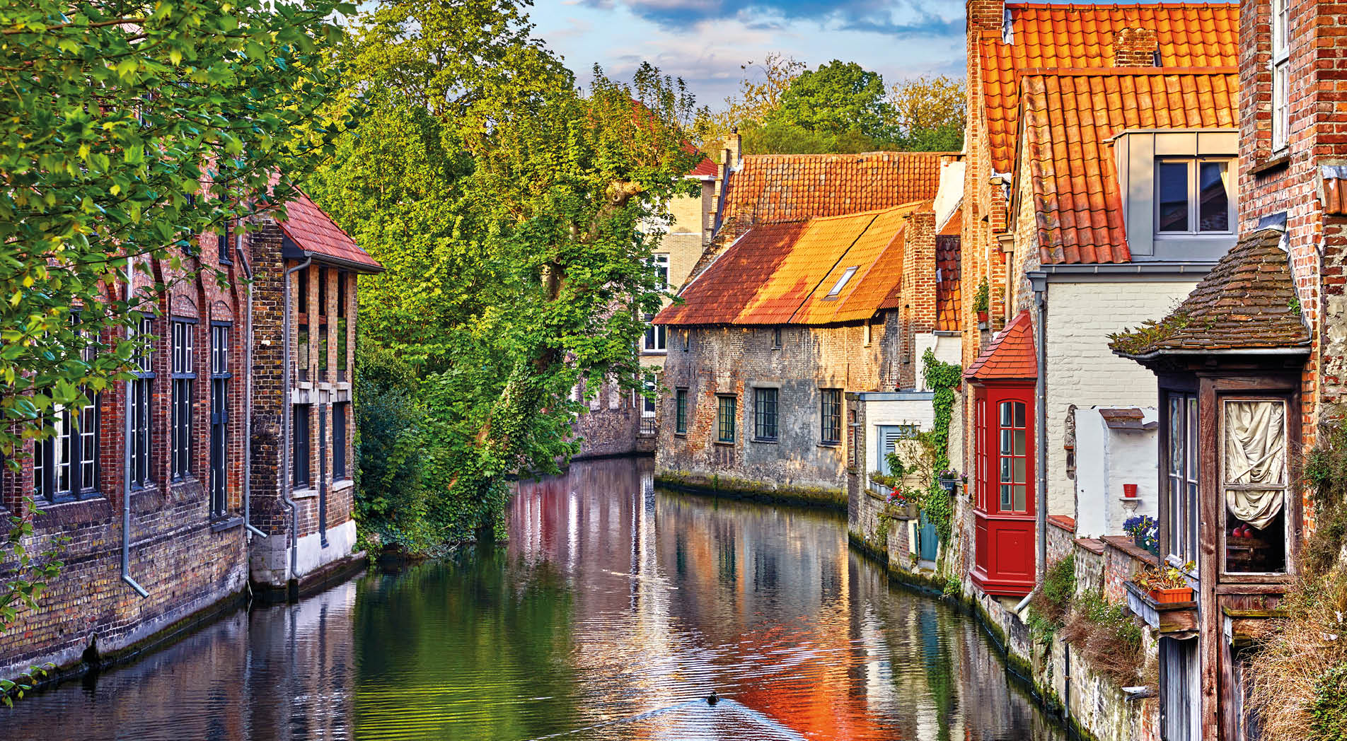 Bruges, Belgium. Medieval ancient houses made of old bricks at water channel with boats in old town. Summer sunset with sunshine and green trees. Picturesque landscape.