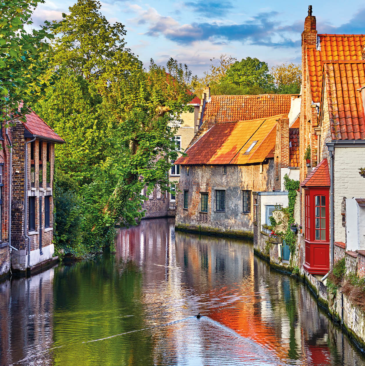 Bruges, Belgium. Medieval ancient houses made of old bricks at water channel with boats in old town. Summer sunset with sunshine and green trees. Picturesque landscape.