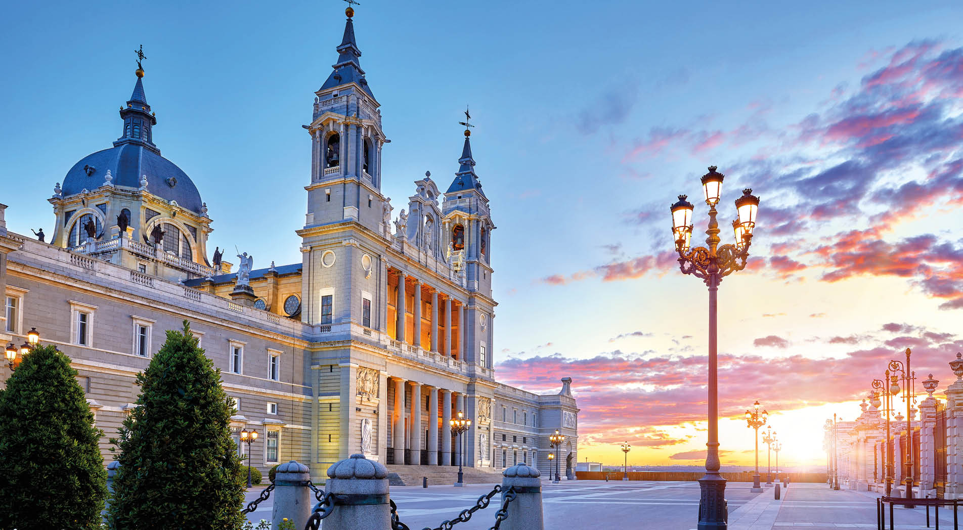 Madrid, Spain. Cathedral Santa Maria la Real de la Almudena at Plaza de la Armeria. Famous landmark with sunset sun, flowers and green bush. Street lamps with illumination and picturesque sky