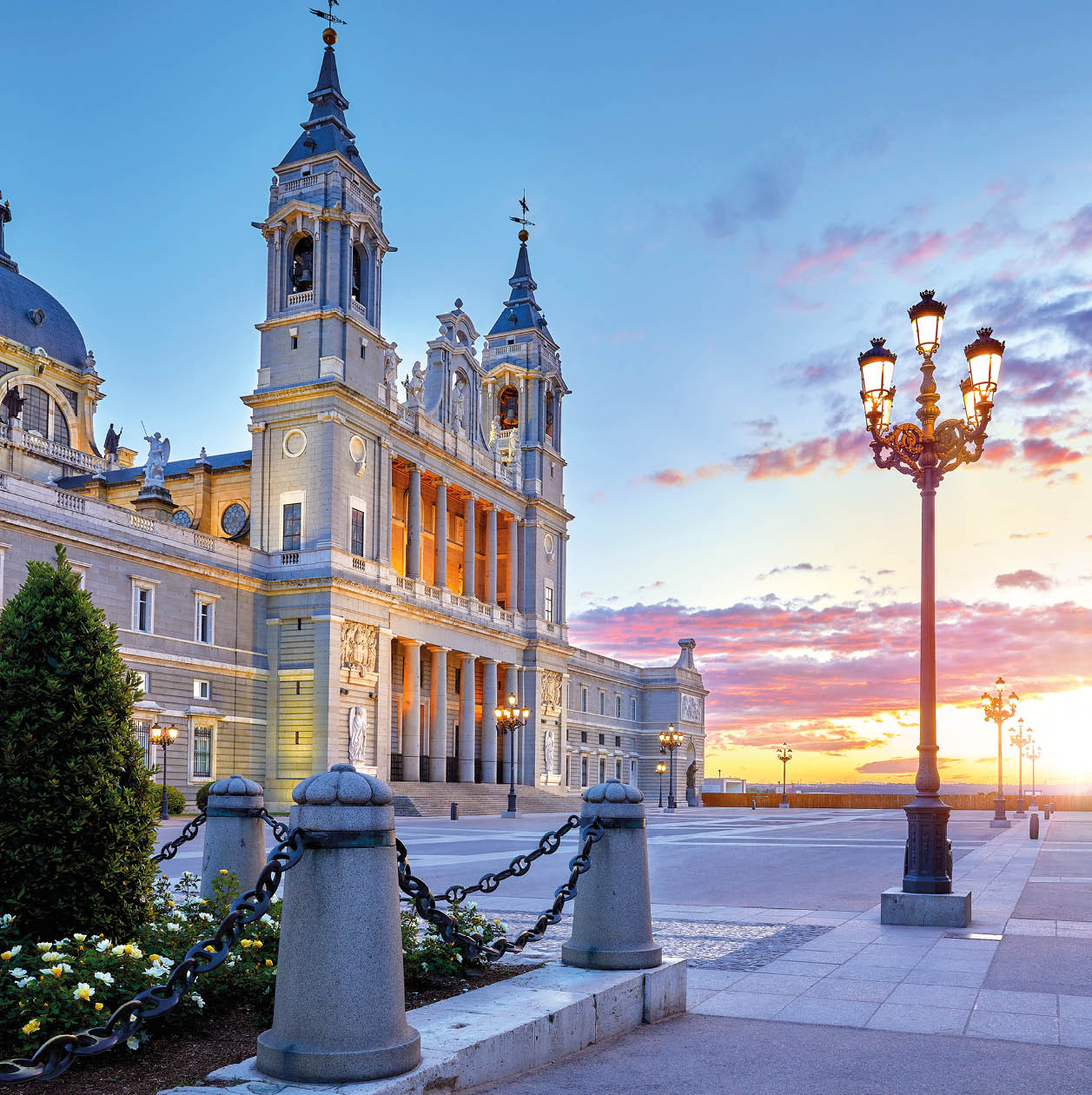 Madrid, Spain. Cathedral Santa Maria la Real de la Almudena at Plaza de la Armeria. Famous landmark with sunset sun, flowers and green bush. Street lamps with illumination and picturesque sky